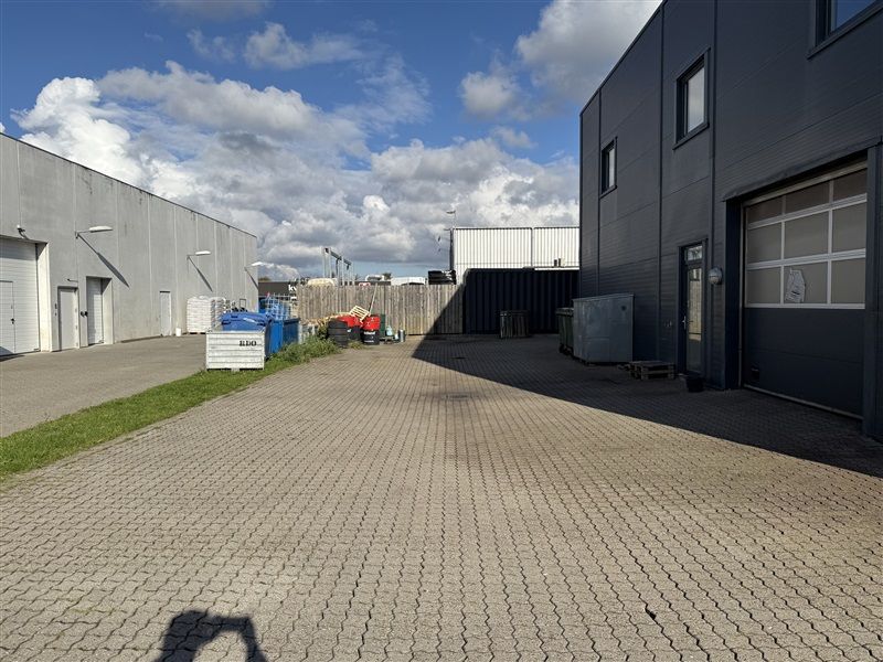 Ude er en stor parkeringsplads med flere garager og bygninger i baggrunden Lejlighedsbygningen er stærkt skyggelagt og der