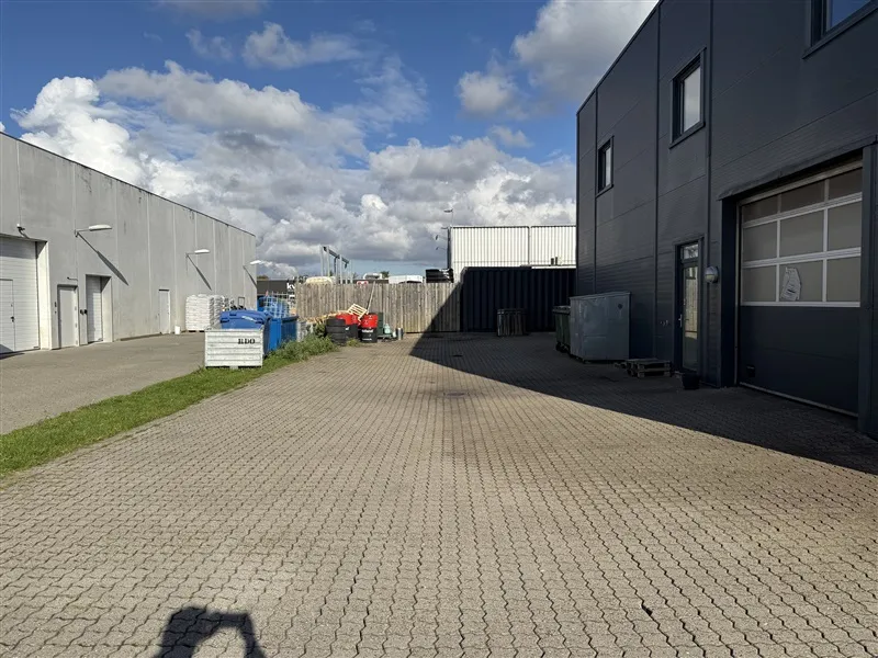 Ude er en stor parkeringsplads med flere garager og bygninger i baggrunden Lejlighedsbygningen er stærkt skyggelagt og der