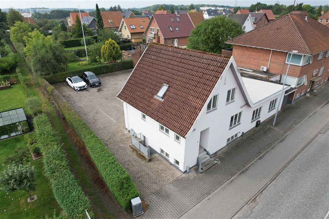 Luftfoto af et hvidt hus med brun tagdækning og garage samt to parkerede biler og grønne haver og stier