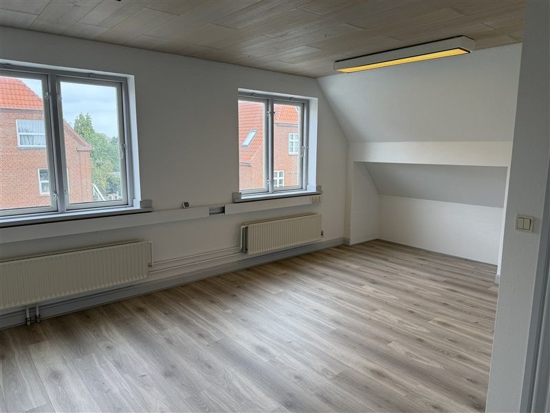 Et kontorlokal med to store vinduer og lysegult lys på loftet. Under vinduet er der radiatorer og gulvtæppe.