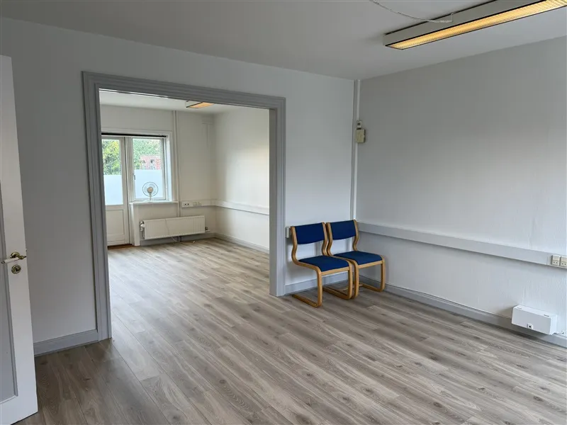 En tom og ren kontorrum med hvide vægge og gulv. To blå stole står mod væggen. En tærskel og dør ligger mod venstre. To