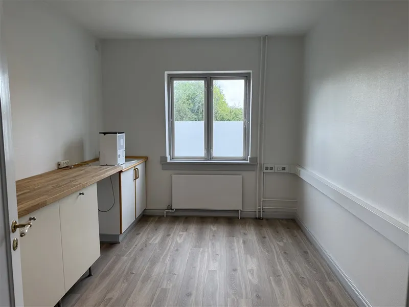 En tom, hvidmalet kontorværelse med trægulv og et vindue med udsigt til park.