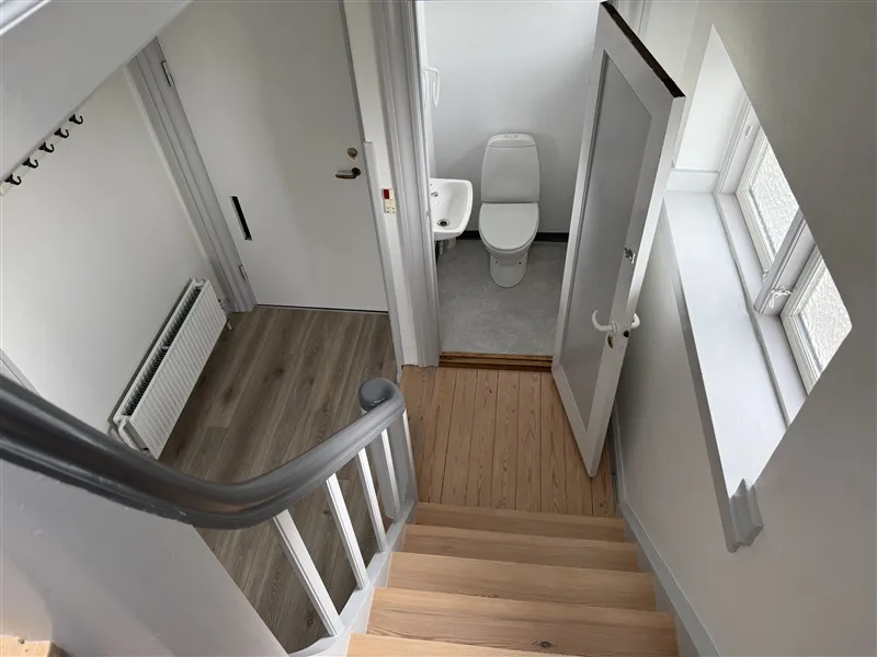 Trapper med lyse trætrin og metalgevær fører til et badeværelse med hvid toilet og hvidt badekar.