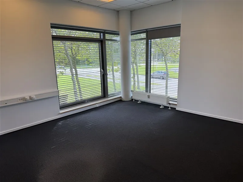 Lokale med store vinduer og mørk gulvbelægning. Her er en radiator og stikkontakter. Udenfor går en bil langs en asfaltvej.
