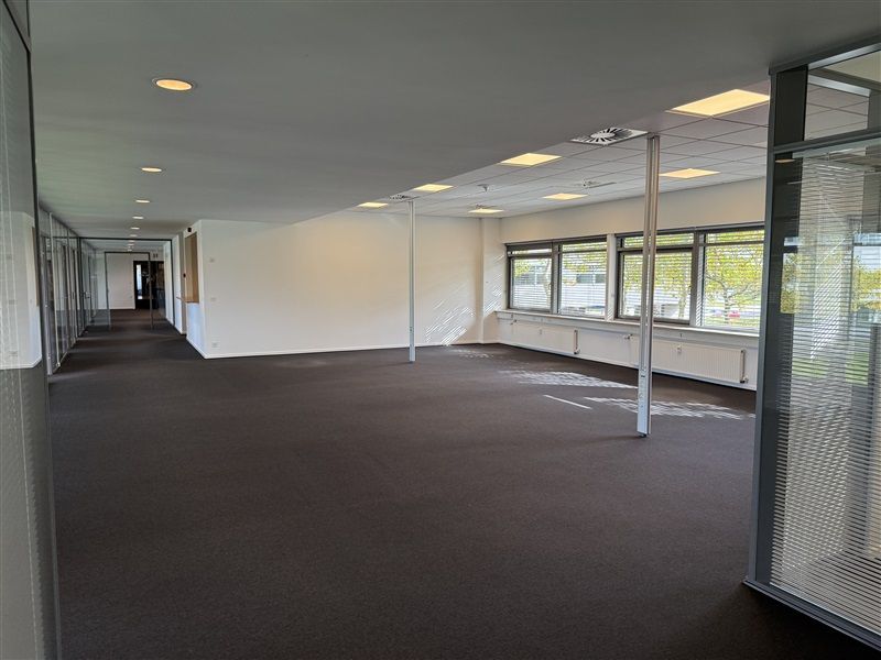 Et stort, lyse og tomt kontorlokale med lysegule vægge og sort gulv. Værelset har store glasruder og lysekroner i loftet.