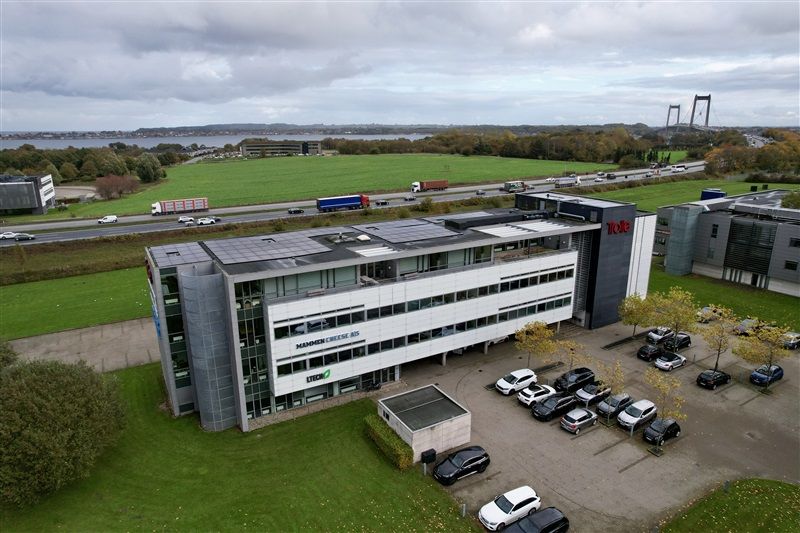 Fabrikbygning i Fredericia med solfanger og biler parkeret udenfor, baggrund med skov, vand og bro