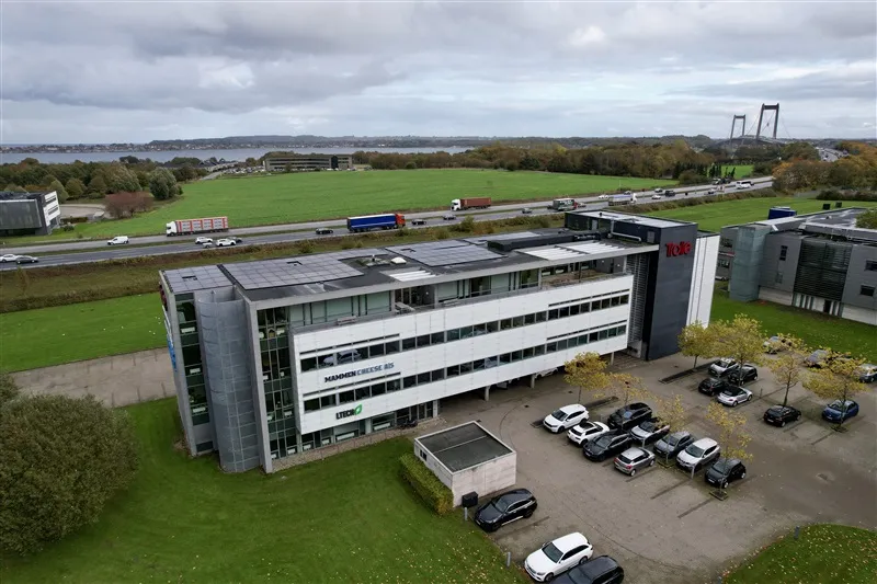 Fabrikbygning i Fredericia med solfanger og biler parkeret udenfor, baggrund med skov, vand og bro