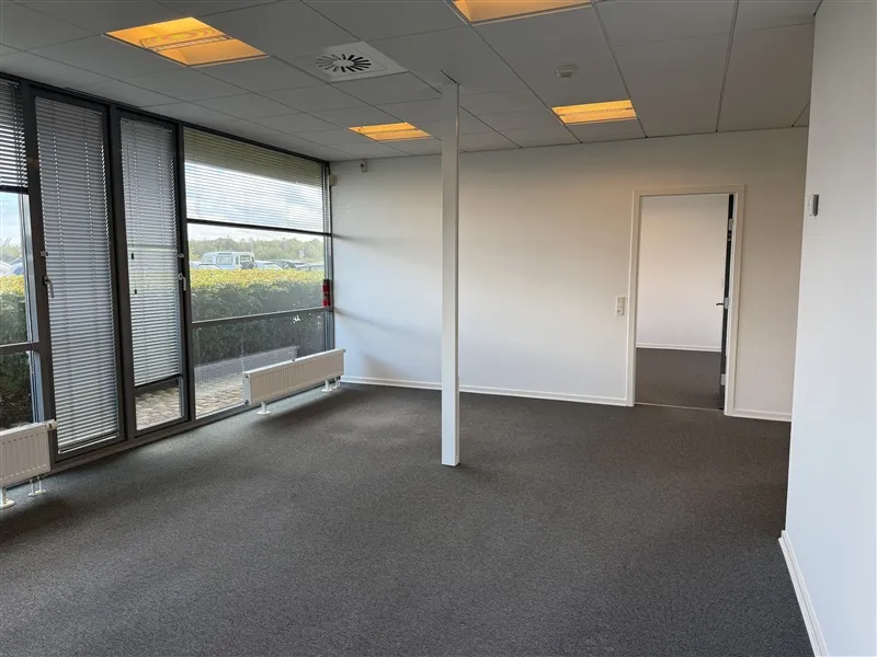 Et tomt kontor med store vinduer, hvide vægge, en hvid dør og gulvbelægning af mørk grå farve.