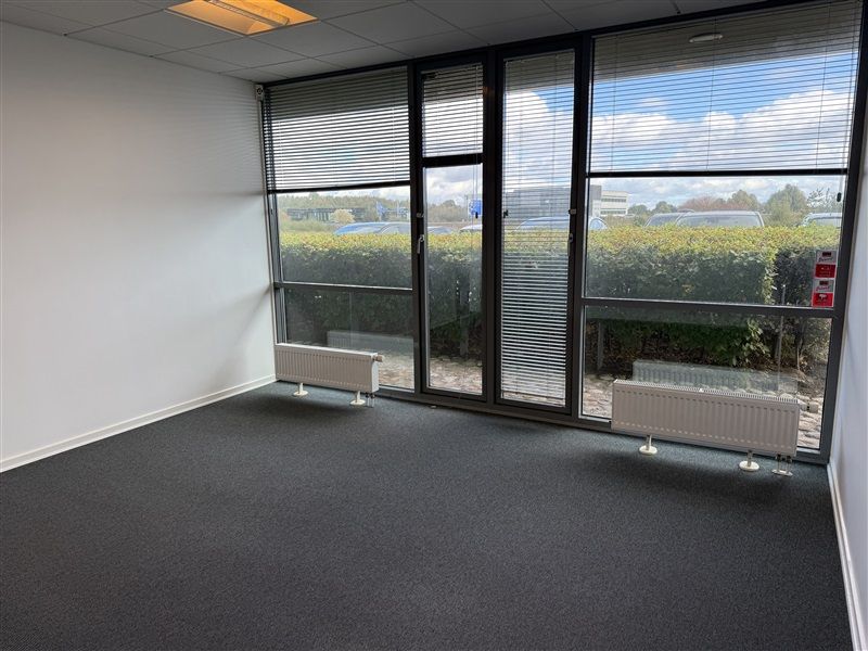 Et tomt rum med hvide vægge, gulvtæppe, radiatorer og glasdøre, der giver udsigt til en parkeringsplads med biler og planter