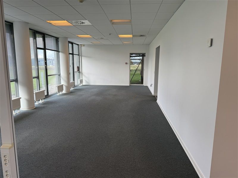 Et tomt kontorrum med lysegult gulv og hvide vægge. Der er flere store ruder med gardiner og flere klimaanlæg i hjørnerne.