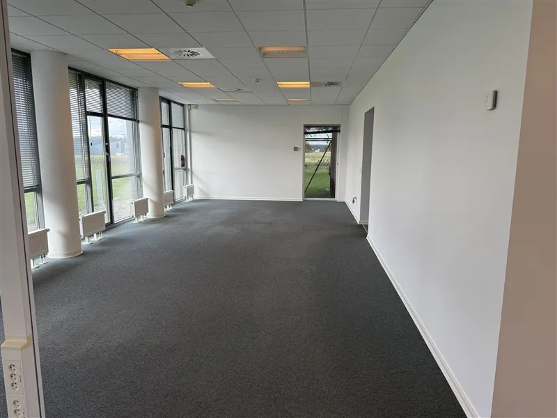 Et tomt kontorrum med lysegult gulv og hvide vægge. Der er flere store ruder med gardiner og flere klimaanlæg i hjørnerne.