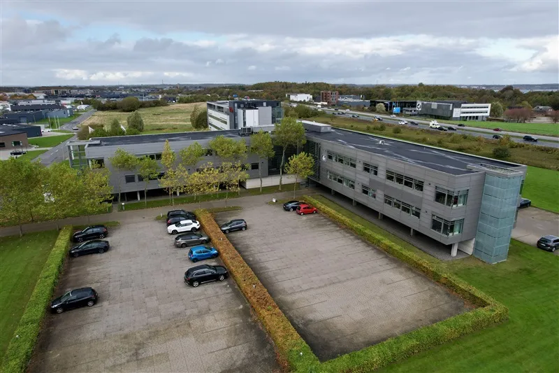 Et kontorlejemål med en stor parkeringsplads og en række biler parkeret. Bygningen er større og placeret midt på vejen.