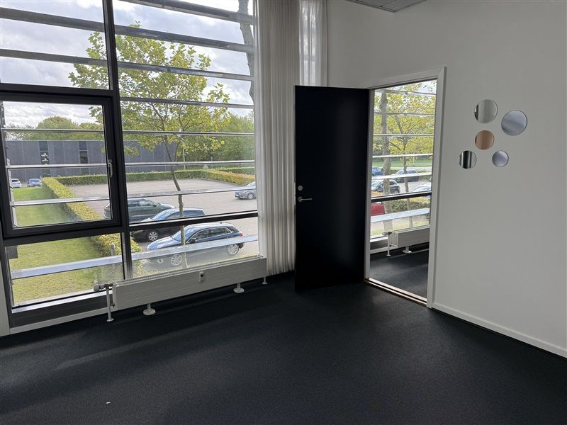 Kontorlokaler med store ruder og parkeringspladser udenfor. En dør er åben og reflekterer det omgivende område.