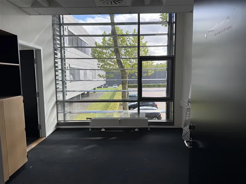 Et lyst og rummeligt kontorlokale med lysevinduer og smukke udsigt til parkeringspladsen med grønne træer og flere biler