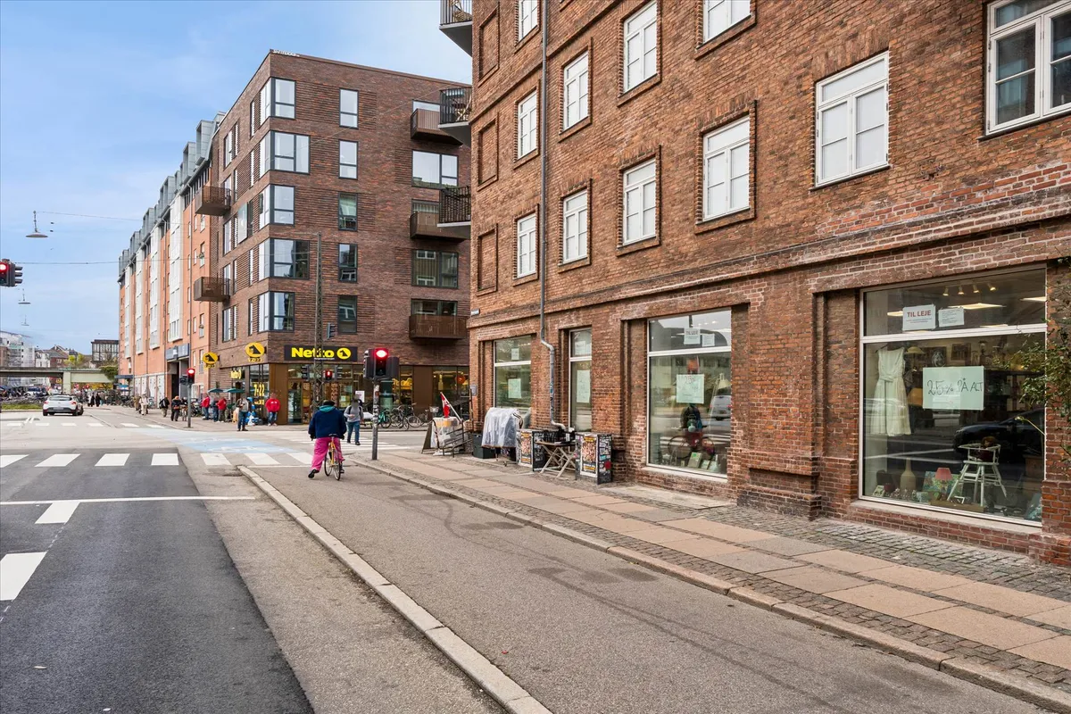 Butik på Mimersgade og Borgmestervangen. Her er søjler og gadeviske, hvor folk går og cykler. Et rødt lys blinker og en