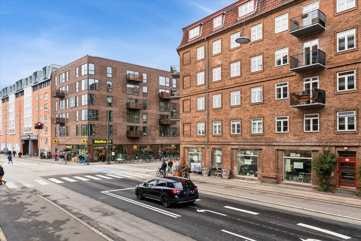 En bil kører på Mimersgade, med bygninger og gadegnæsserne på hver side. Et Netto-butiksskilt er synligt på en af bygningerne