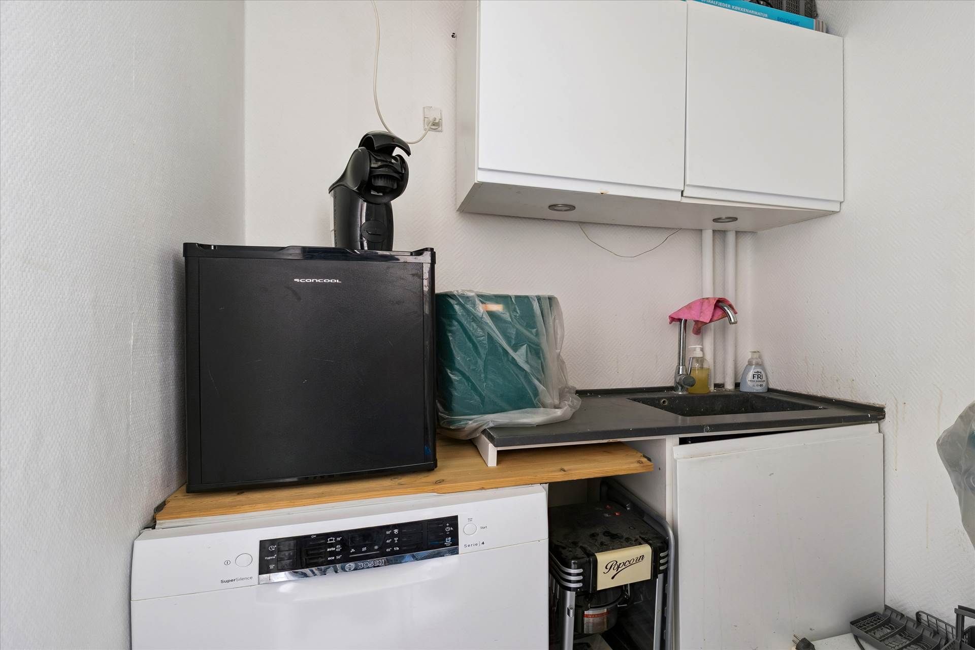 Et lille køkken med en hvid skab, en mikrobølgeovn, en tørretumbler og en skraldpose samt en stribet stol og en skraldespand.