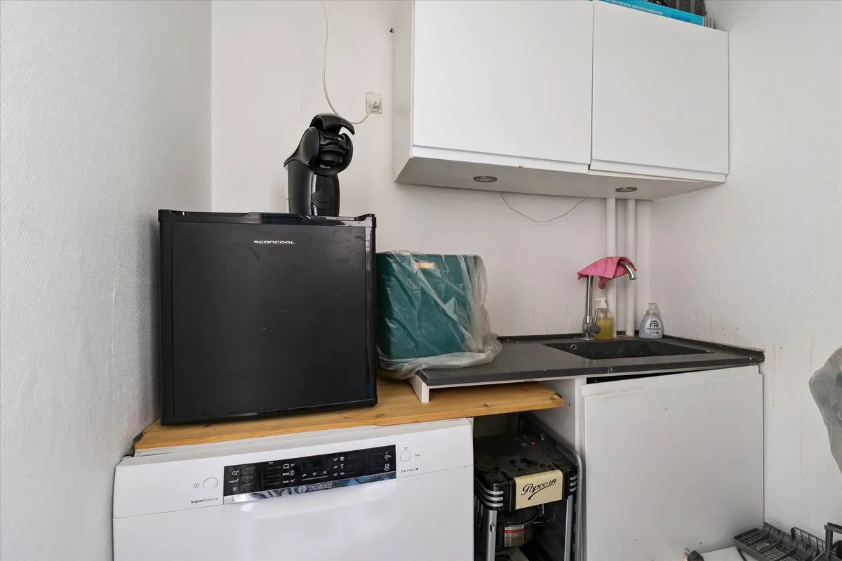 Et lille køkken med en hvid skab, en mikrobølgeovn, en tørretumbler og en skraldpose samt en stribet stol og en skraldespand.