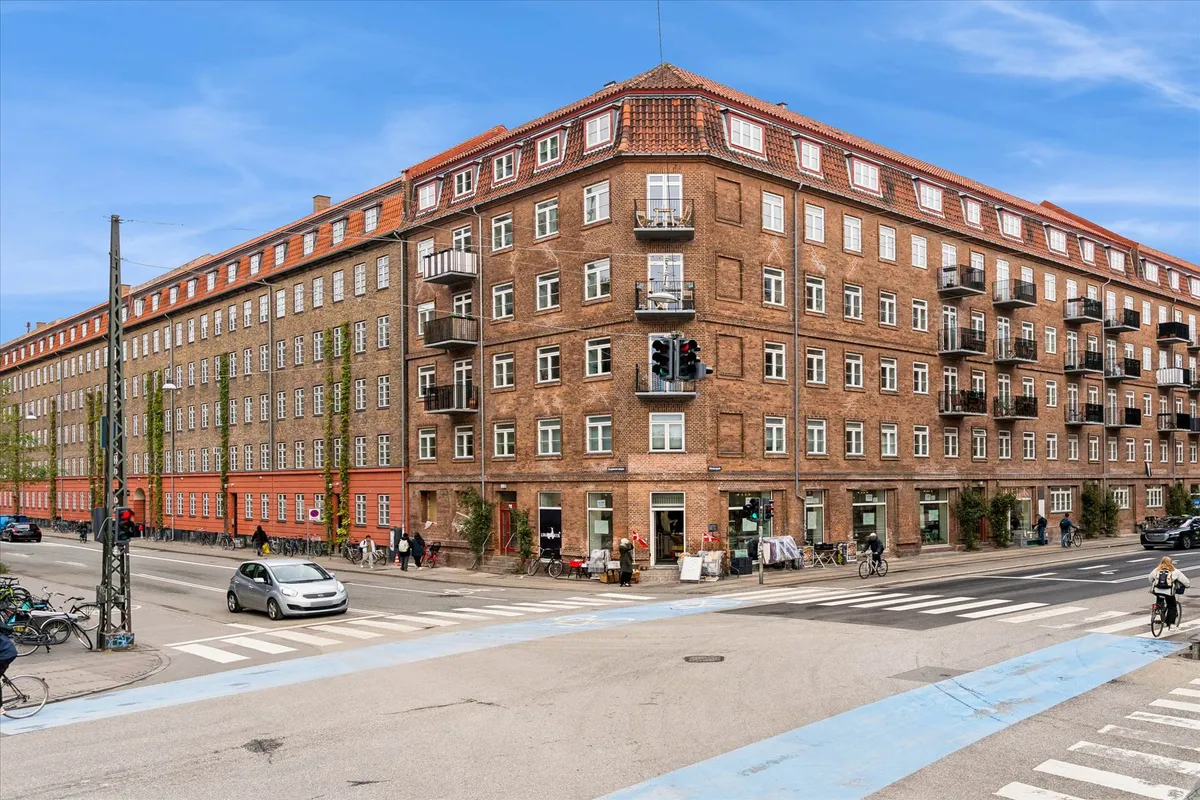 Mennesker går på gaden og cykler ved hjørnebygningen med mange lejligheder og hjørnebutikker, hvor trafikken er tæt på.