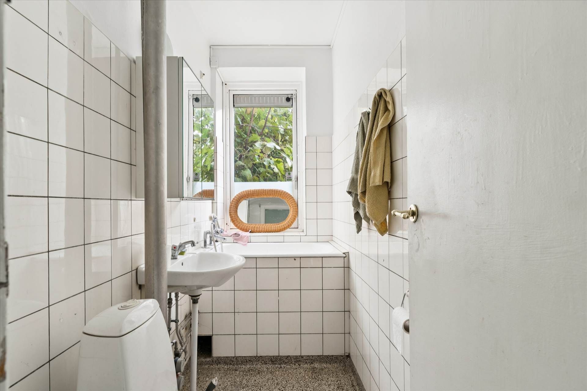 Et lille badeværelse med hvide kakeloverflader, to toiletter, et vindue og et håndklæde hængende på væggen