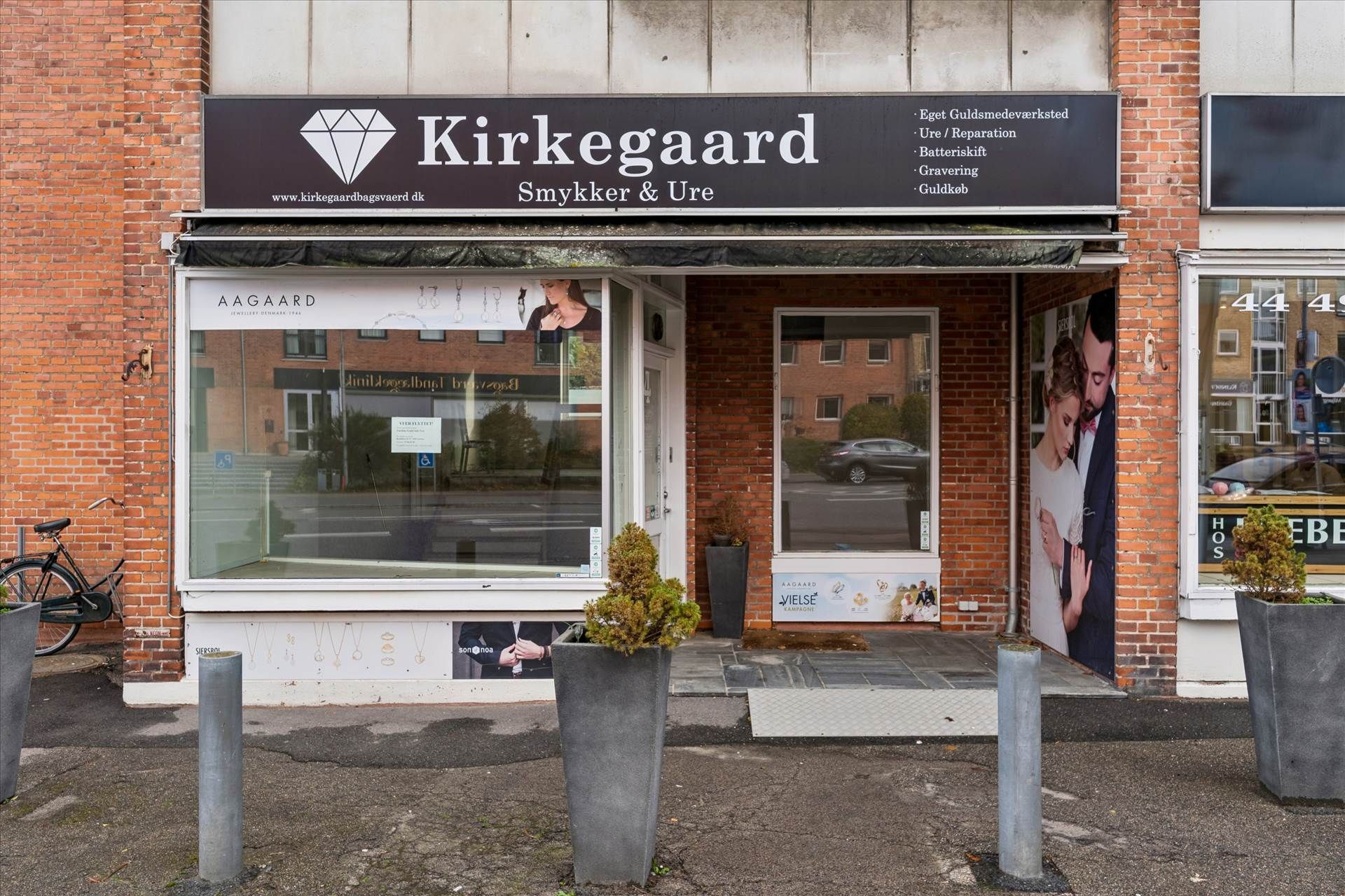 Forretningslokal med udstillingsvindue og tekst Kirkegaard. Glasskærm med reklame og tekst Aagaard. To potteplanter og tekst