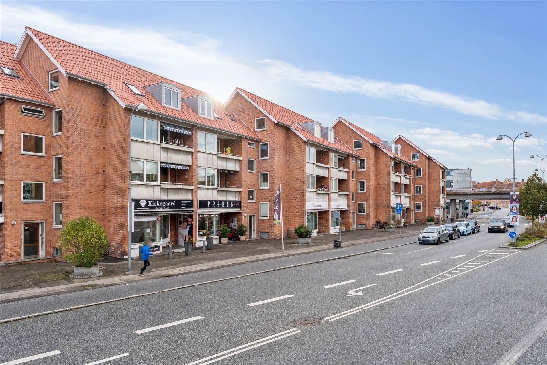 Butik i 1. sal med udgang til gågade med mange biler parkeret. En person i blåt tøj går ud af butikken. Solskin og skyer i