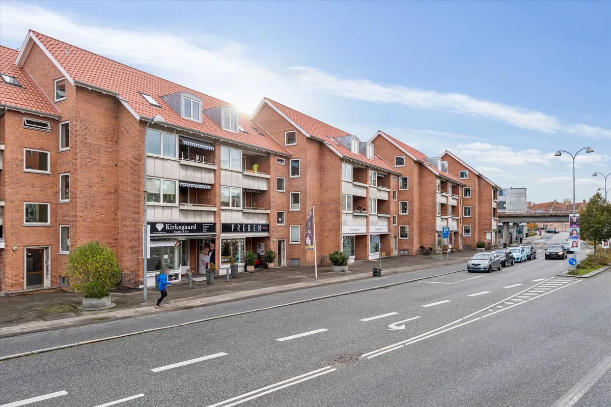 Butik i 1. sal med udgang til gågade med mange biler parkeret. En person i blåt tøj går ud af butikken. Solskin og skyer i