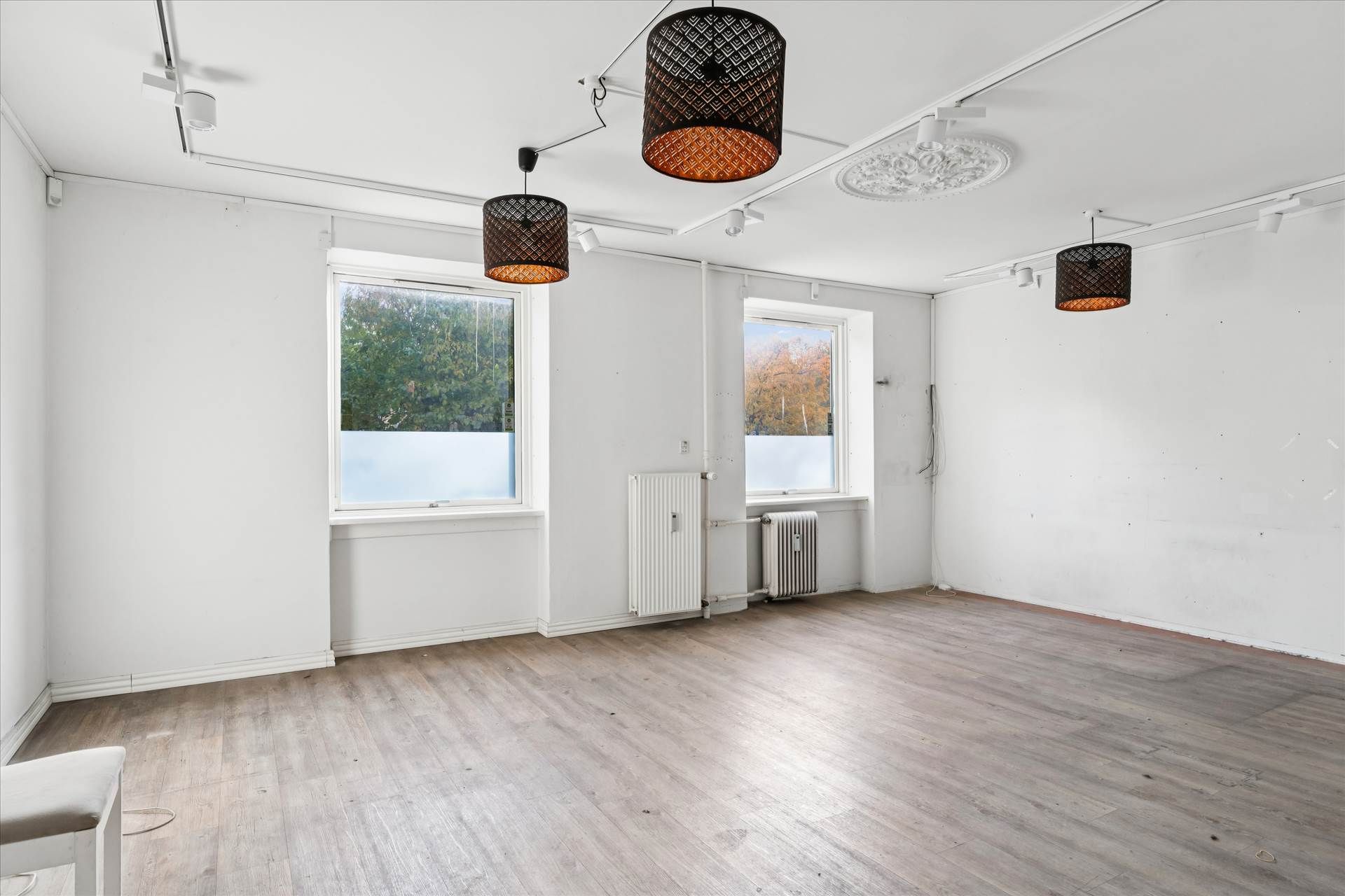 Et lyst og indbydende rum med hvide vægge, to store vinduer, tre pendantlys og hårdt trægulv. Værelset er ubenyttet og der