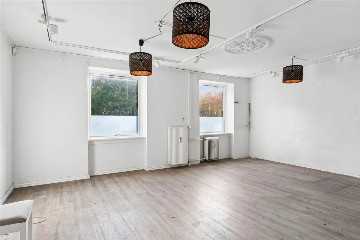 Et lyst og indbydende rum med hvide vægge, to store vinduer, tre pendantlys og hårdt trægulv. Værelset er ubenyttet og der