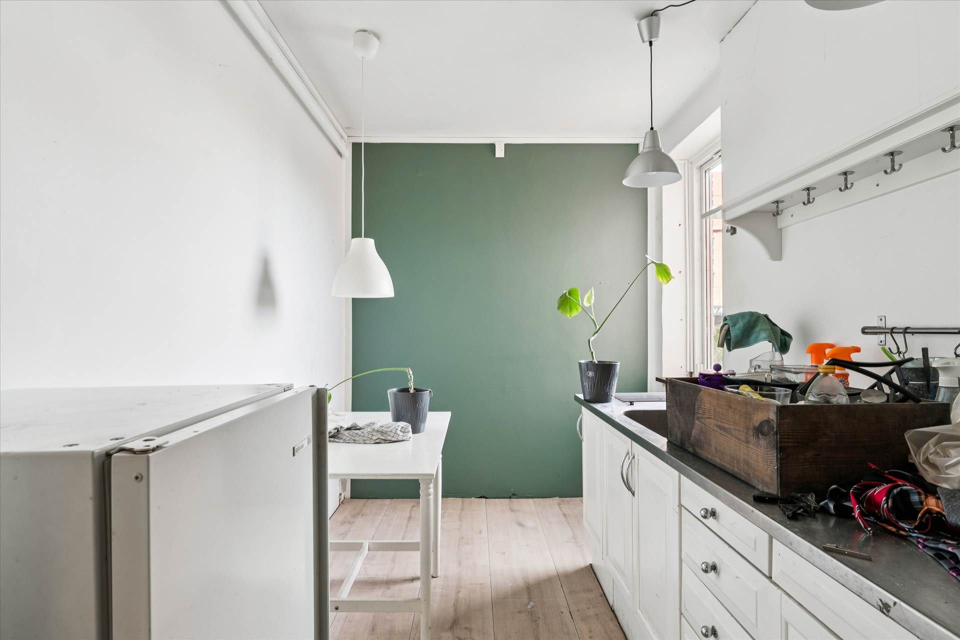 Simpel og pæn køkken med køkkenbord og plante, to hængelampe, en køleskab og en trækasse med diverse genstande på tælleren