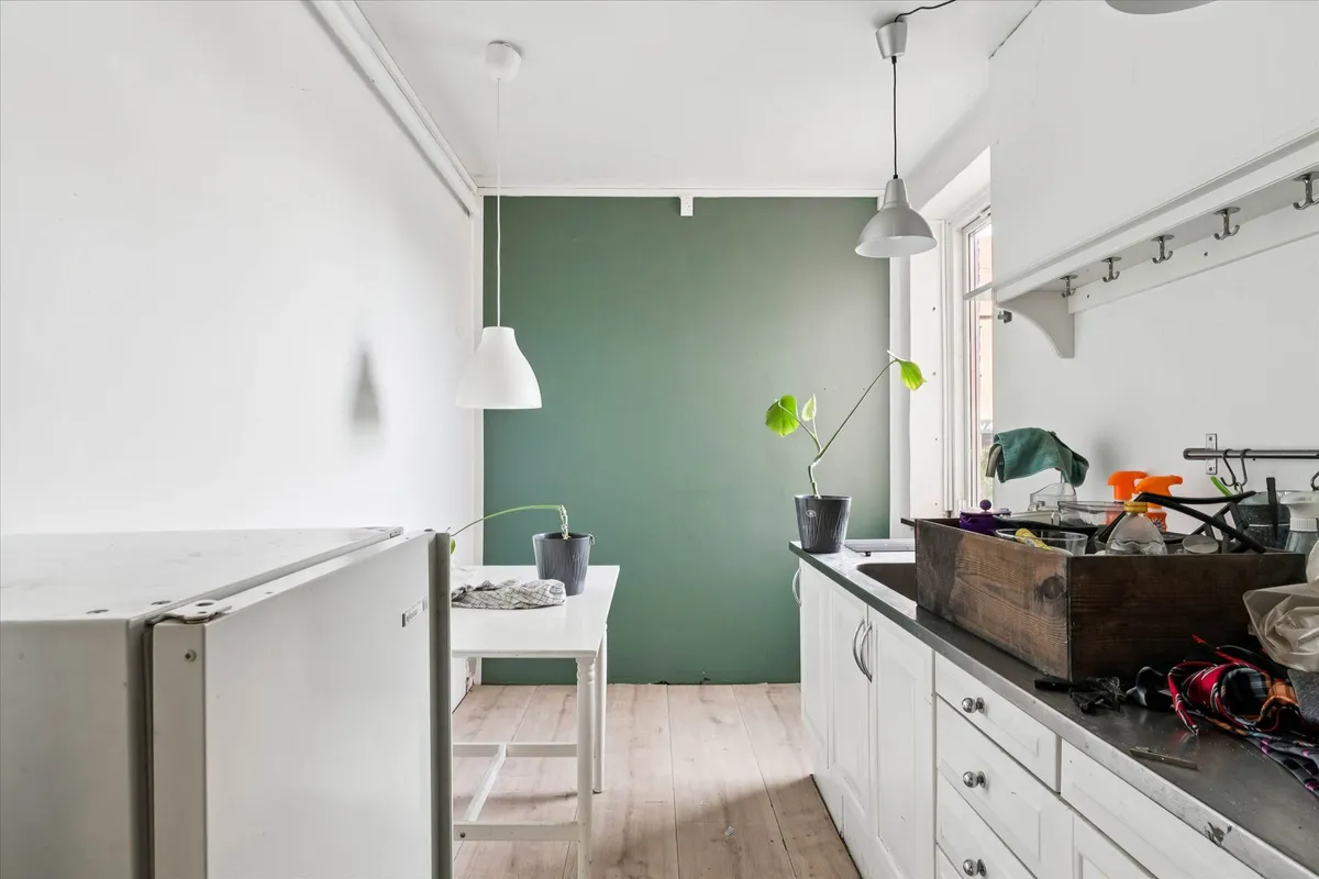 Simpel og pæn køkken med køkkenbord og plante, to hængelampe, en køleskab og en trækasse med diverse genstande på tælleren