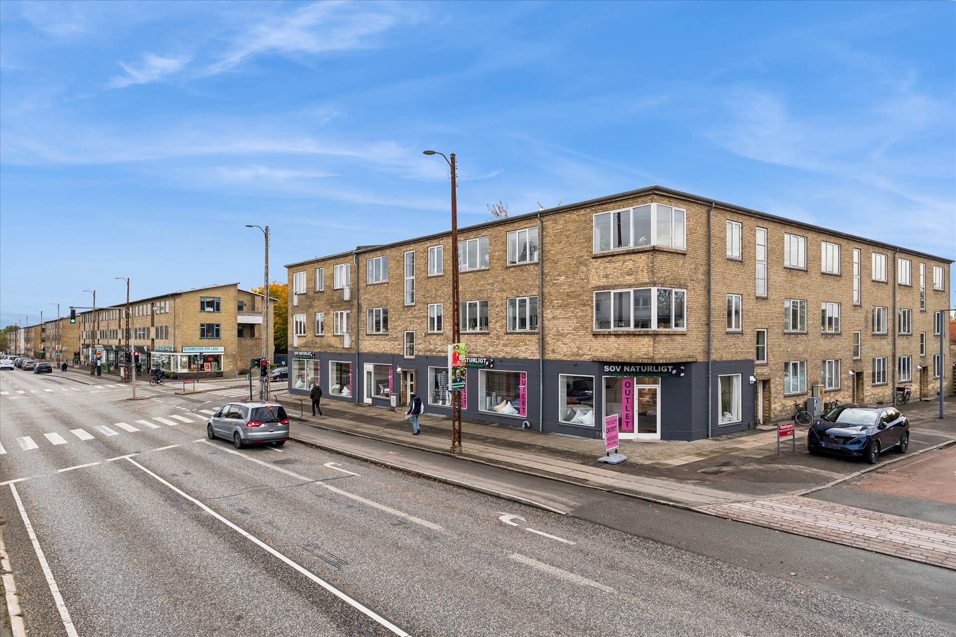 En stor bygning med mange vinduer og butikker på gadehjørnet af Roskildevej. Biler parkeret og folk vandrer forbi.