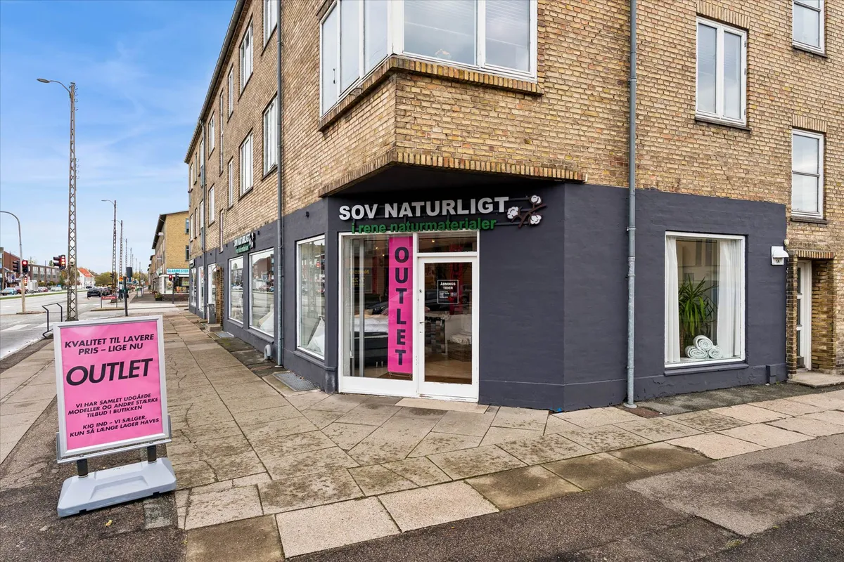 Outlet butik med udsolgte varer og nylige lagersalg. Pink skilt med tekst på dansk. Lys blå bygning med vinduer og røde