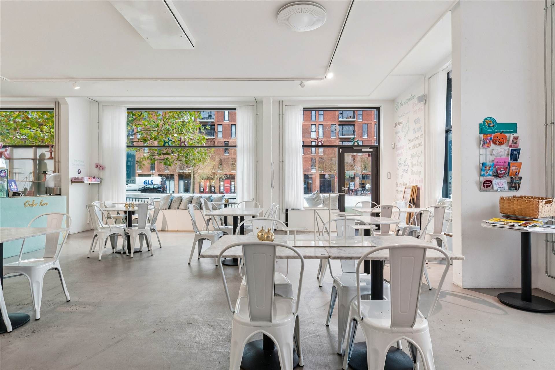 Moderne café med hvide møbler og en stor glasdør. Væggene er hvide med lyse skrivninger. I vinduet ser man træer og en