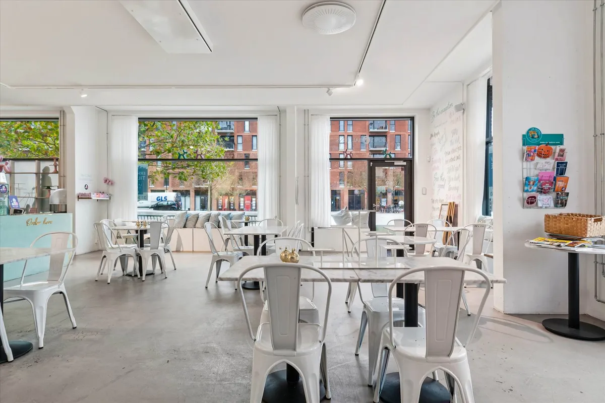 Moderne café med hvide møbler og en stor glasdør. Væggene er hvide med lyse skrivninger. I vinduet ser man træer og en