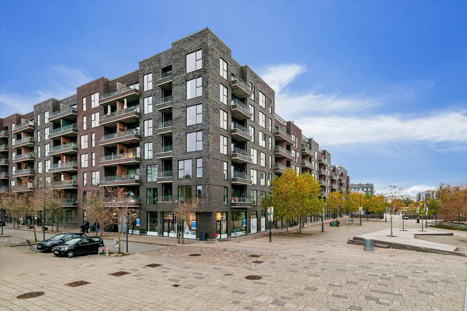 En stor højhuslejlighed med terrasser og udsigt til byen i baggrunden. En større gågade med parkeringsplads og butikker.