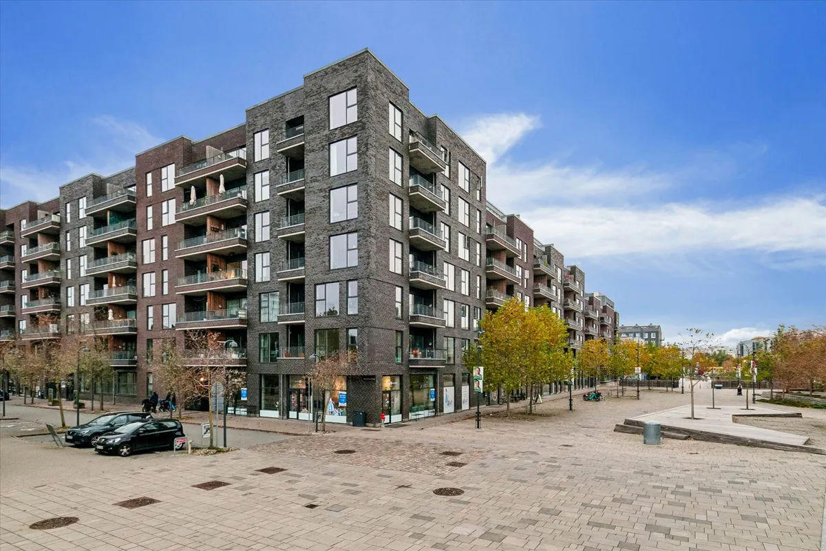 En stor højhuslejlighed med terrasser og udsigt til byen i baggrunden. En større gågade med parkeringsplads og butikker.