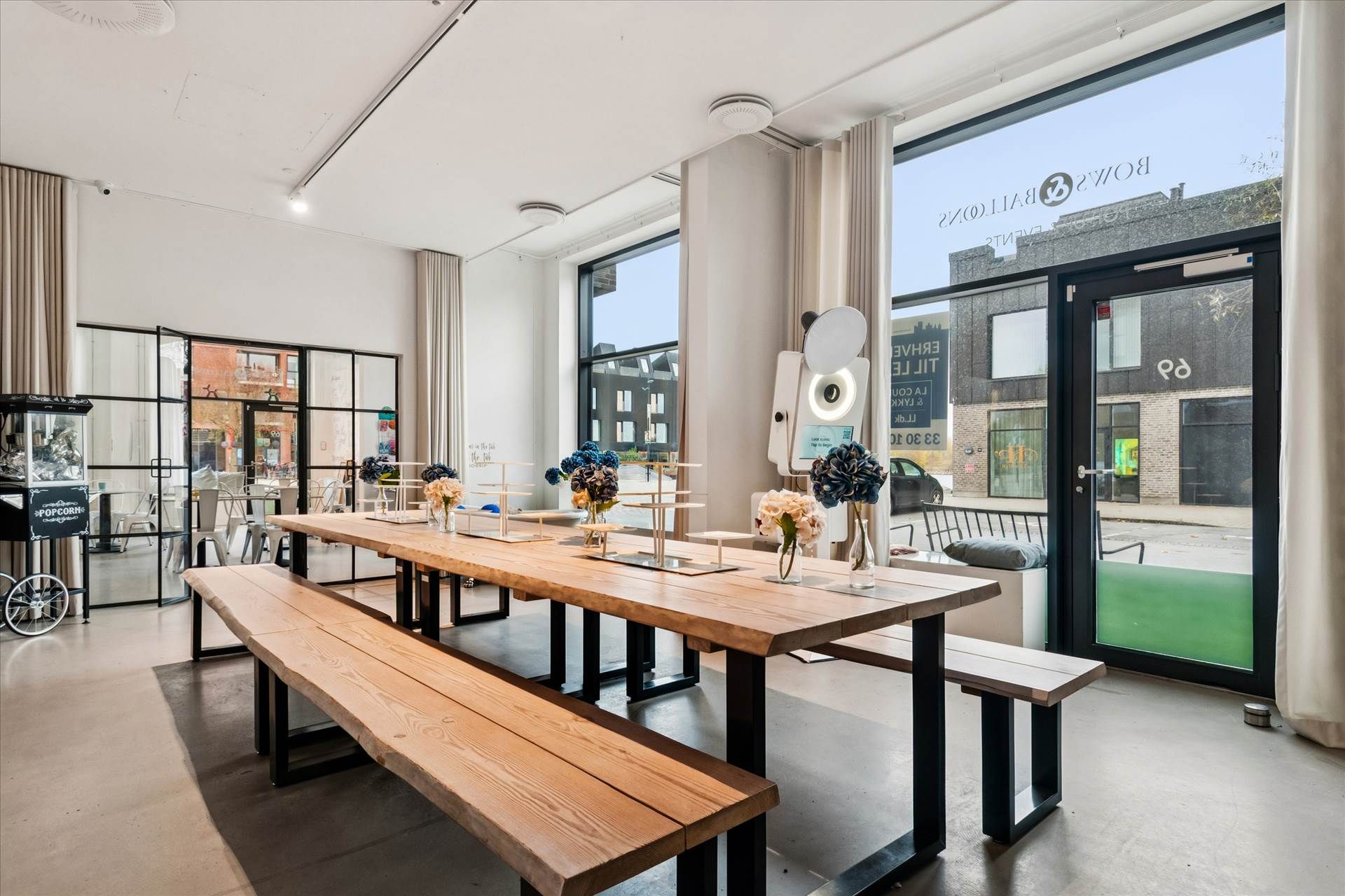 Moderne lokale med to lange træbænke, træbord og blomster, samt store glasruder med udsigt til en bygning og en bil