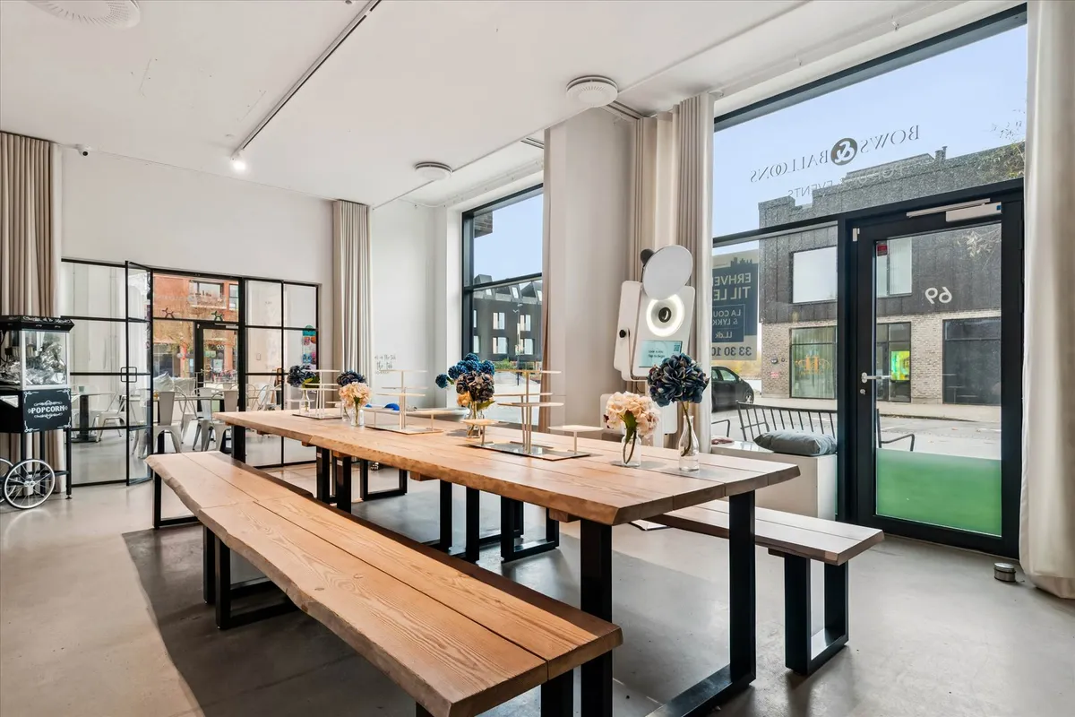 Moderne lokale med to lange træbænke, træbord og blomster, samt store glasruder med udsigt til en bygning og en bil