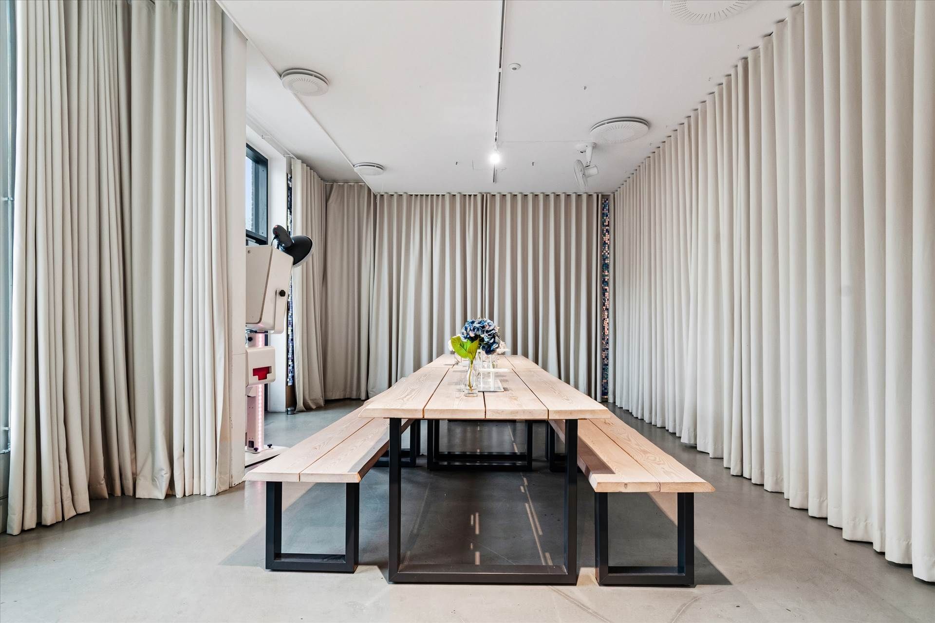 En stor træbord med to bænke og en vase med blå blomster står i et rum med hvide væggardøjer og loftsventilatorer