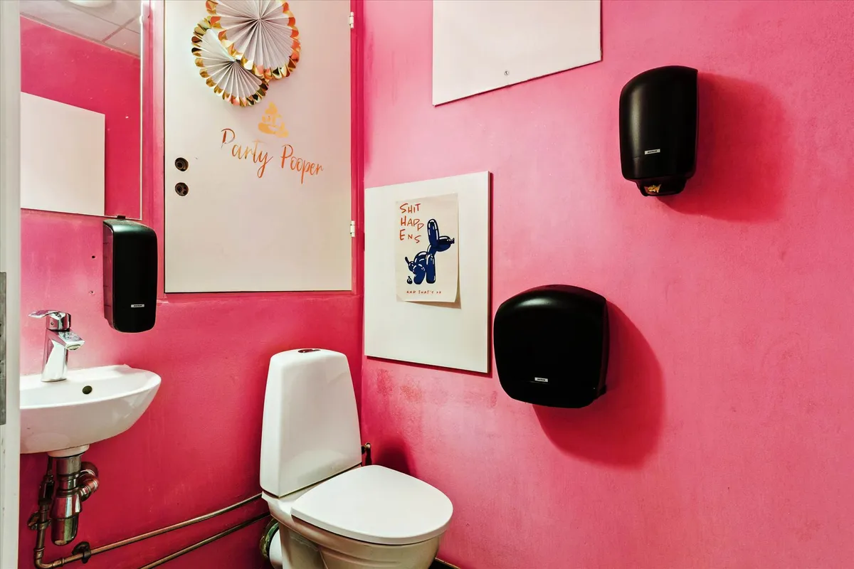 Privat toilet med pink vægge, hvid toilet, hængende toiletpapir, håndvask med vandhane og et papir med tekst og tegning på