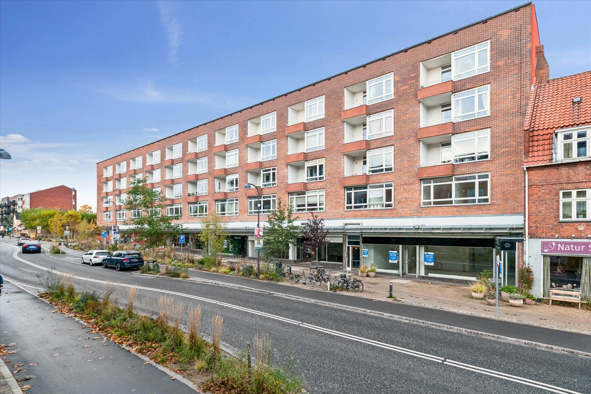 Butik med facade af røde mursten og glasvinduer. Foran er der parkerede biler og cykler. Der er en skilt med teksten Natur