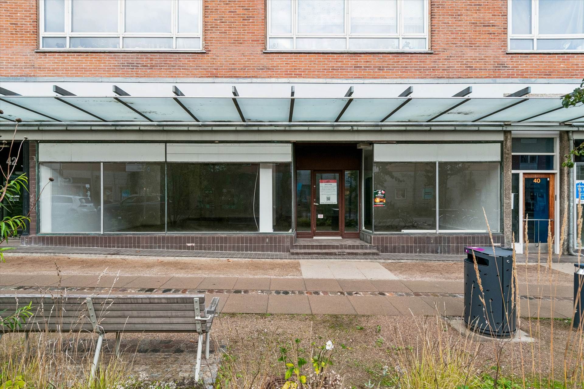 Et lukket butikslokal med store ruder, brun dør og nummer 40. En glasoverdækning og gul nummerplade foran. To skraldspande