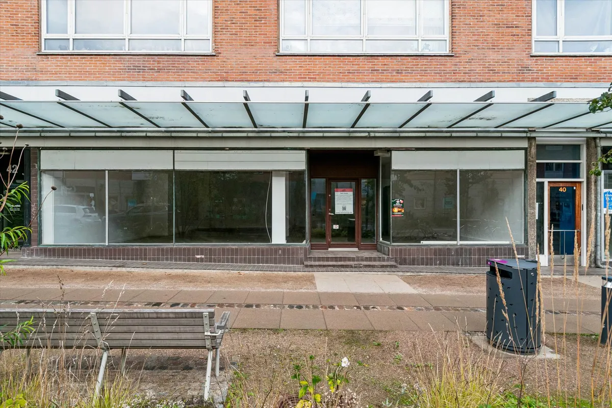 Et lukket butikslokal med store ruder, brun dør og nummer 40. En glasoverdækning og gul nummerplade foran. To skraldspande