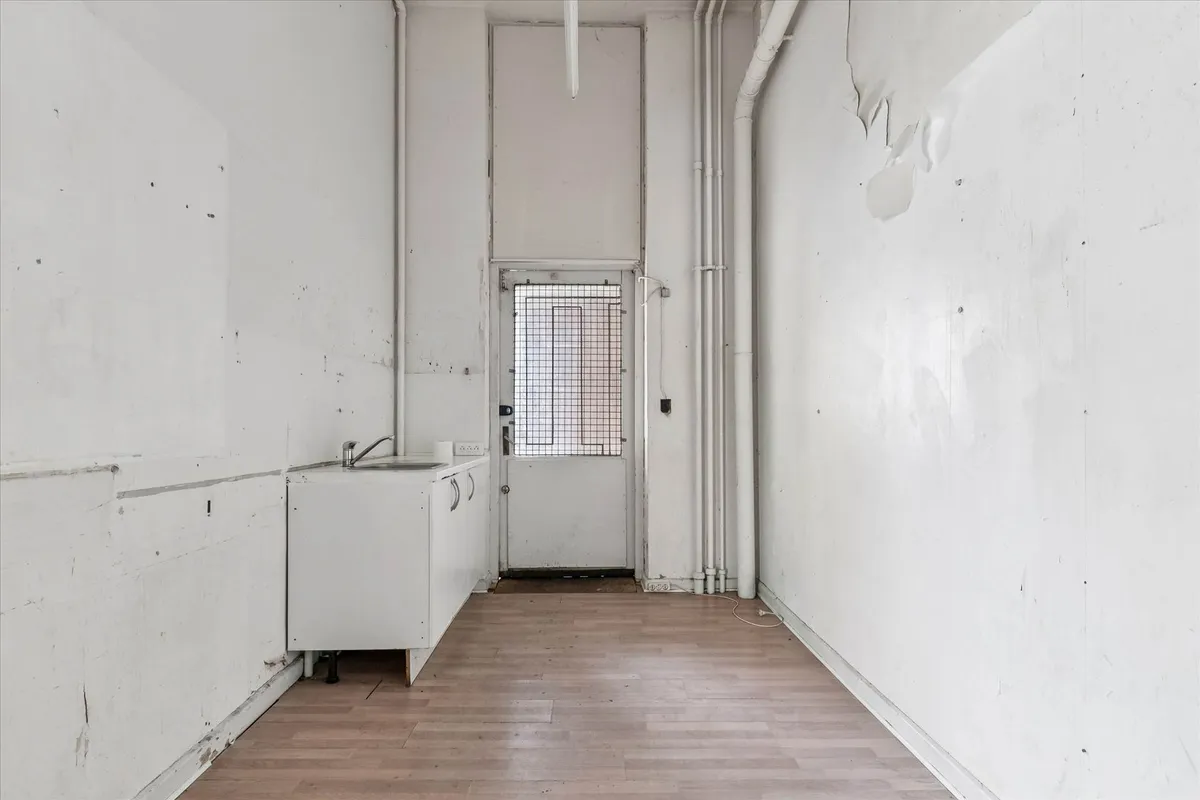 En tom og ren lagerhal med lyse, trægulve, hvide vægge og en simpel, hvid vask samt en lukket dør med et lyskab