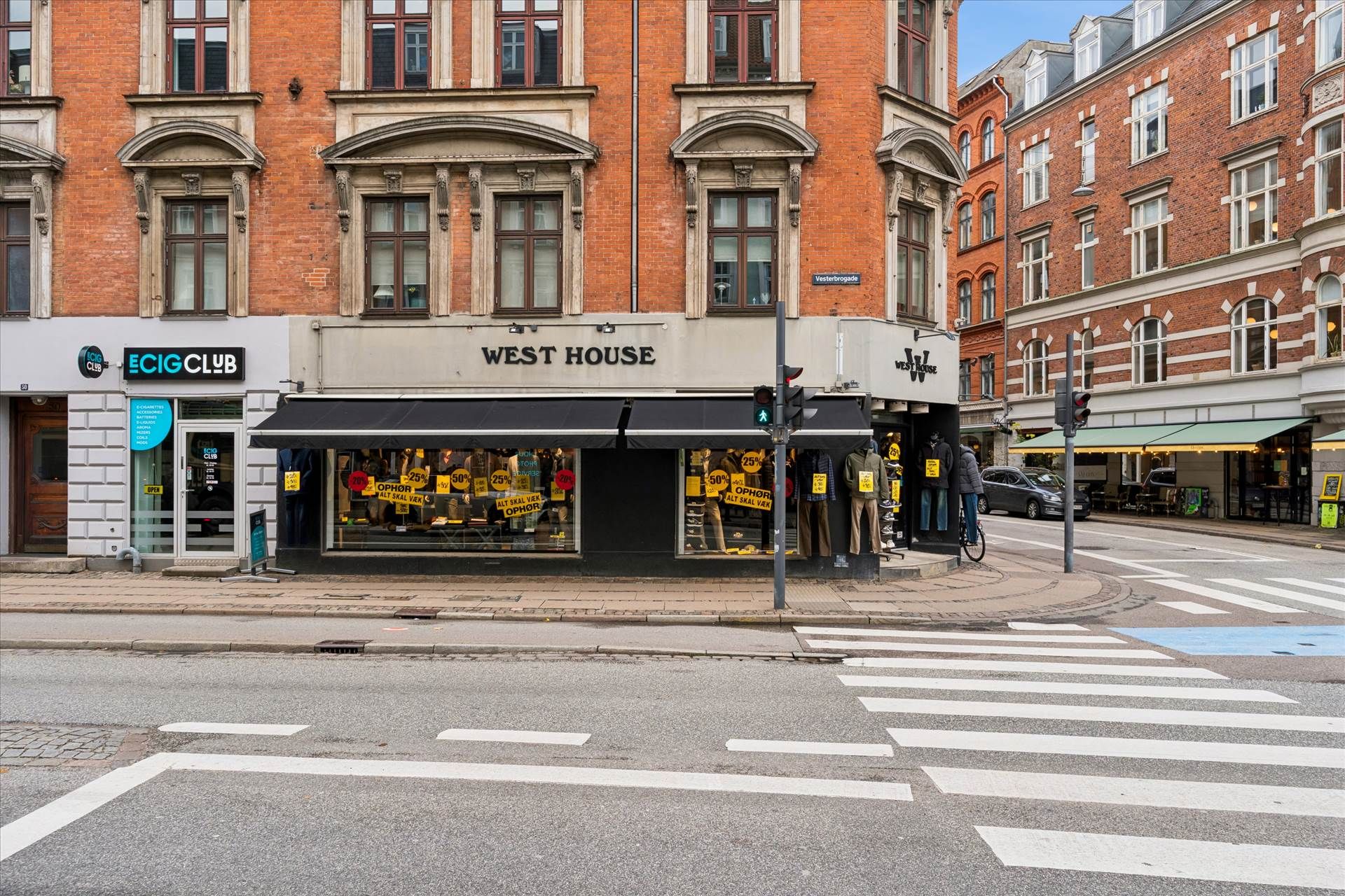 Vesterbro med West House-butik og E-Cig Club-forretning, hvor vejkryds er mærket med hvide streger og en cykel står parkeret