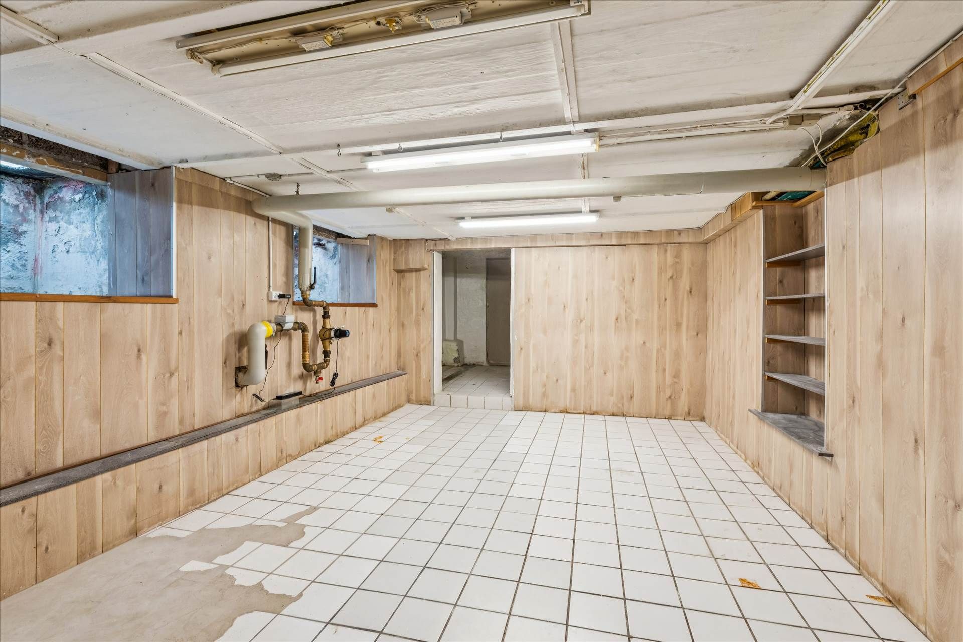 Underjordisk rum med hvide flisegulv, træpaneler og lysekroner samt piper og rør ophængt i væggen.