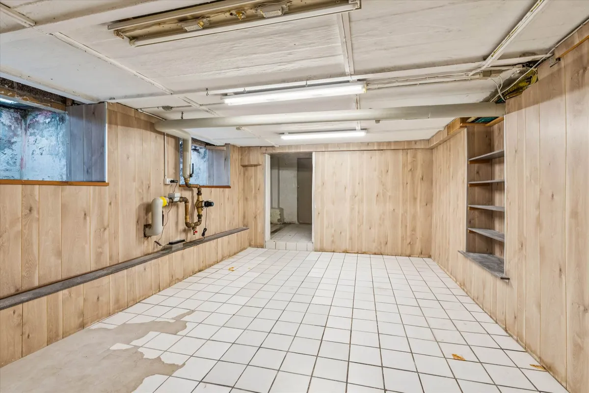 Underjordisk rum med hvide flisegulv, træpaneler og lysekroner samt piper og rør ophængt i væggen.