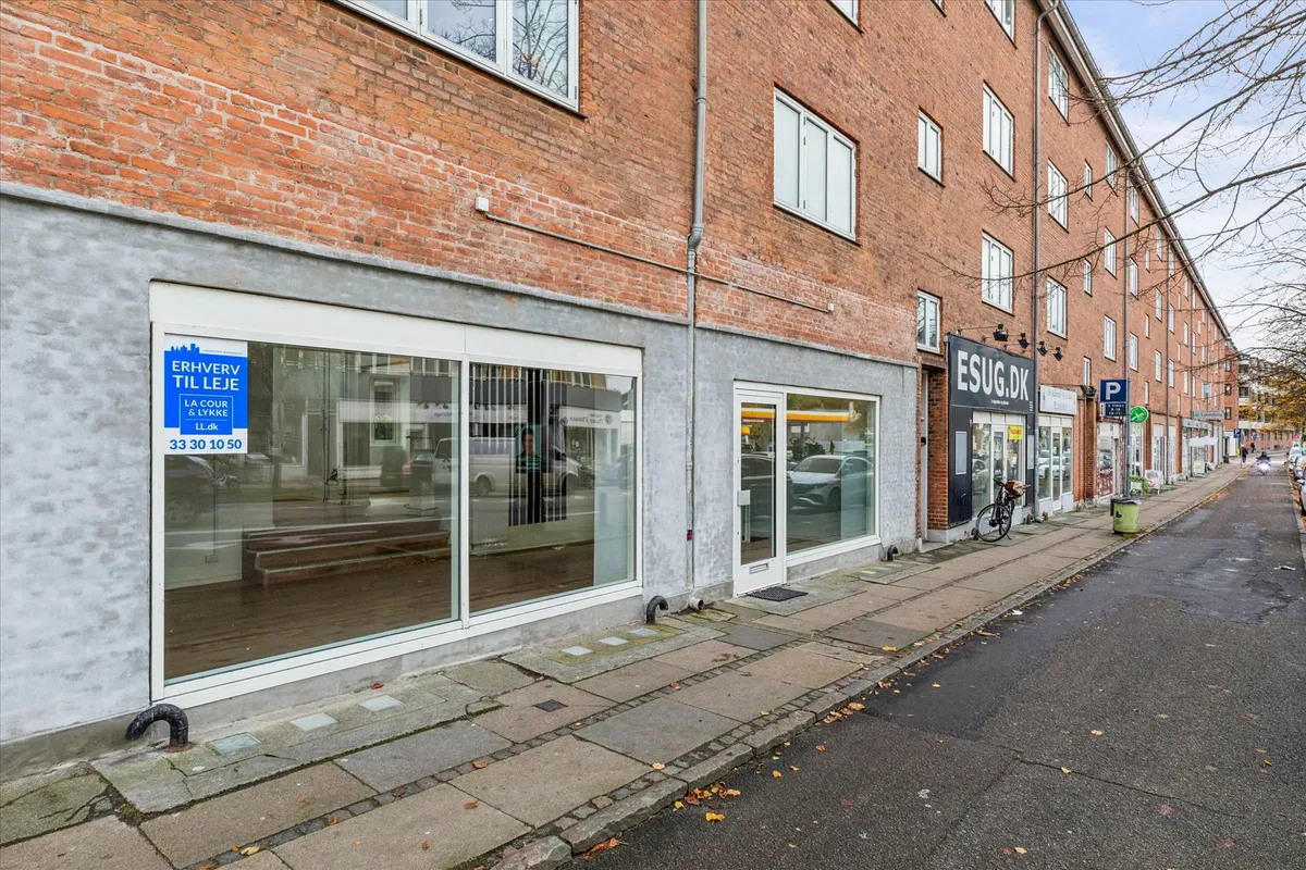 Butik med glasvinduer og skilt med teksten "Erhverv til leje" og telefonnummer 33 30 10 50. Bag butikken er der et skur med