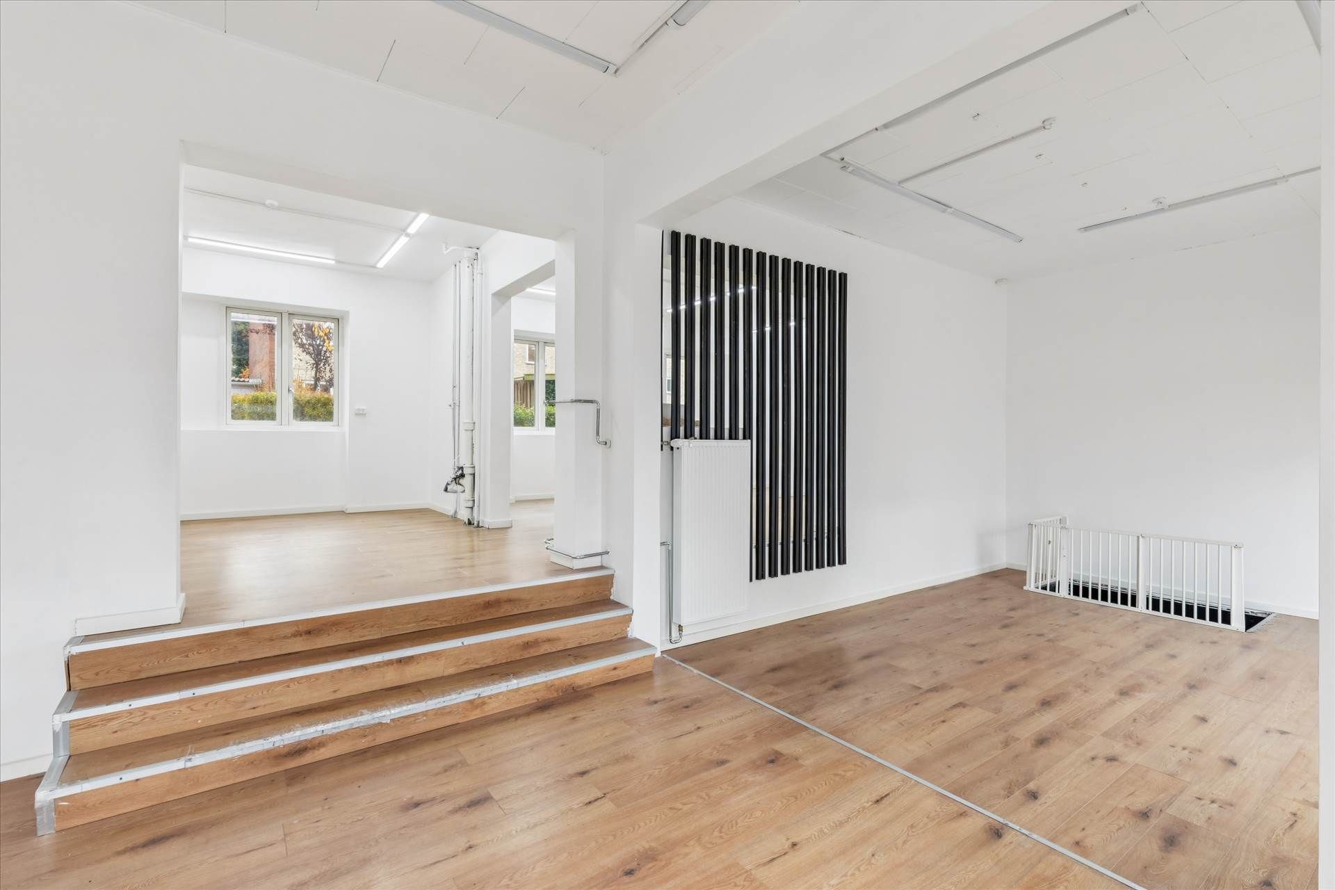 En tom hvid stue med trægulv og trapper til kælderen, samt en hvid spær og strimlet møbel foran en hvid væg